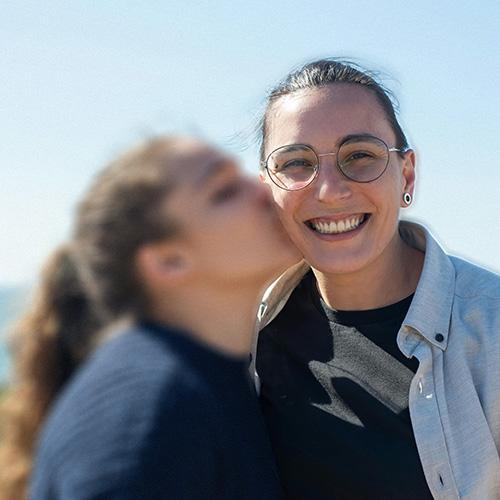 A young female couple, one kissing the other, in front of a blue sky. one woman is blurred and the other one is in full focus. When you move the cursor over the image, the blurred woman comes into focus.